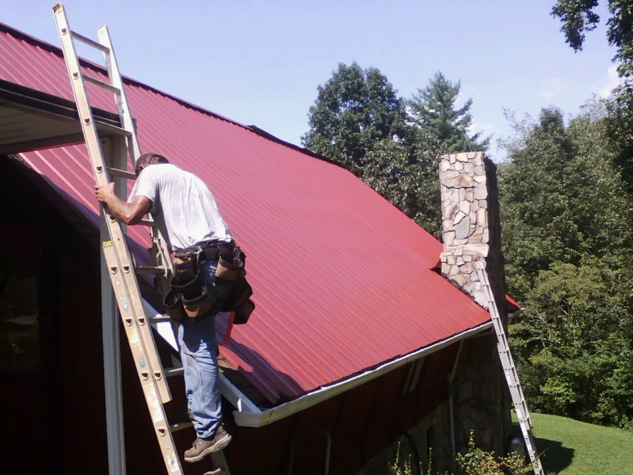 Roofer on a ladder beside a red metal roof and stone chimney — Soffit & Fascia Repair by Roofing Brothers in Dexter